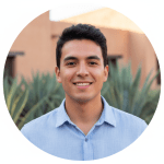 A smiling man wearing a light blue shirt looks at the camera in a circular portrait, with greenery and a building softly blurred in the background.